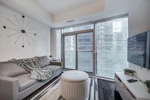Un salon moderne avec un canapé, un pouf et une grande fenêtre donnant sur des immeubles.