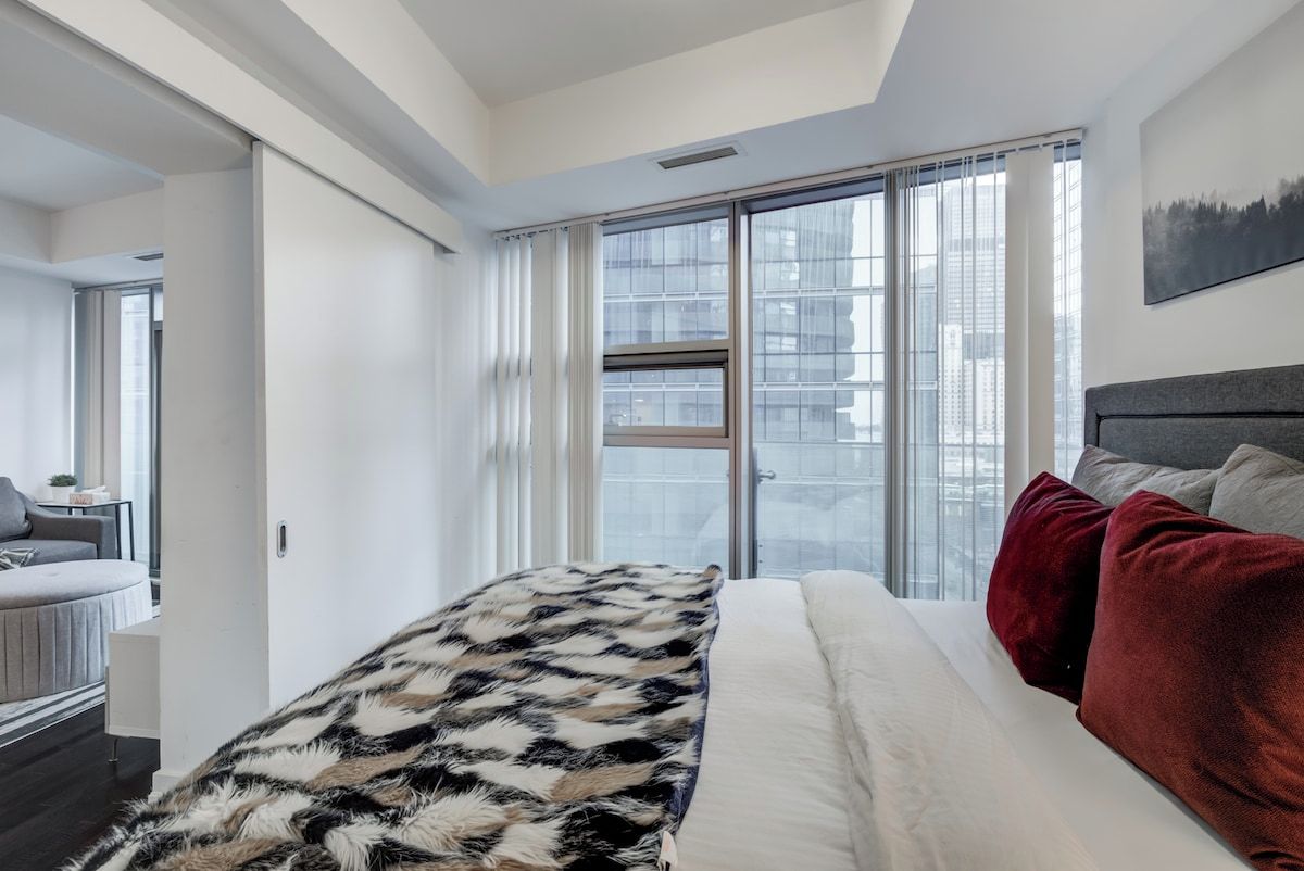 A modern bedroom with a large bed, a view through a window, and a cityscape outside.
