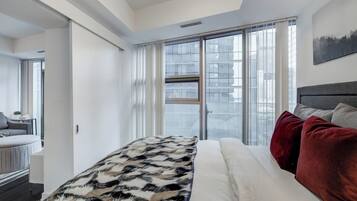 A modern bedroom with a large bed, a view through a window, and a cityscape outside.