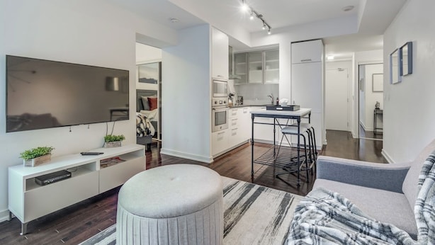 A modern living room with a flat-screen TV, a sofa, a coffee table, and a kitchen area with cabinets and a dining table. - Stylish 1Bed Den Unit in the heart of Dt (Toronto)
