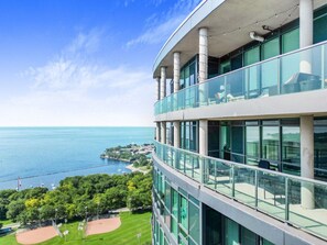 A modern building with a glass facade, a balcony with seating, and a view of the sea and greenery below. - Luxury 2BR Lakeview Penthouse w/ Balcony & Parking (Toronto)