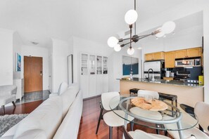 A modern living room with a glass dining table, white sofas, and a kitchen area with wooden cabinets and a microwave. - Luxury 2BR Lakeview Penthouse w/ Balcony & Parking (Toronto)