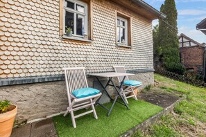 A house with a unique fish-scale patterned exterior wall, a small patio with two chairs and a table, and a garden area with grass and a potted plant.