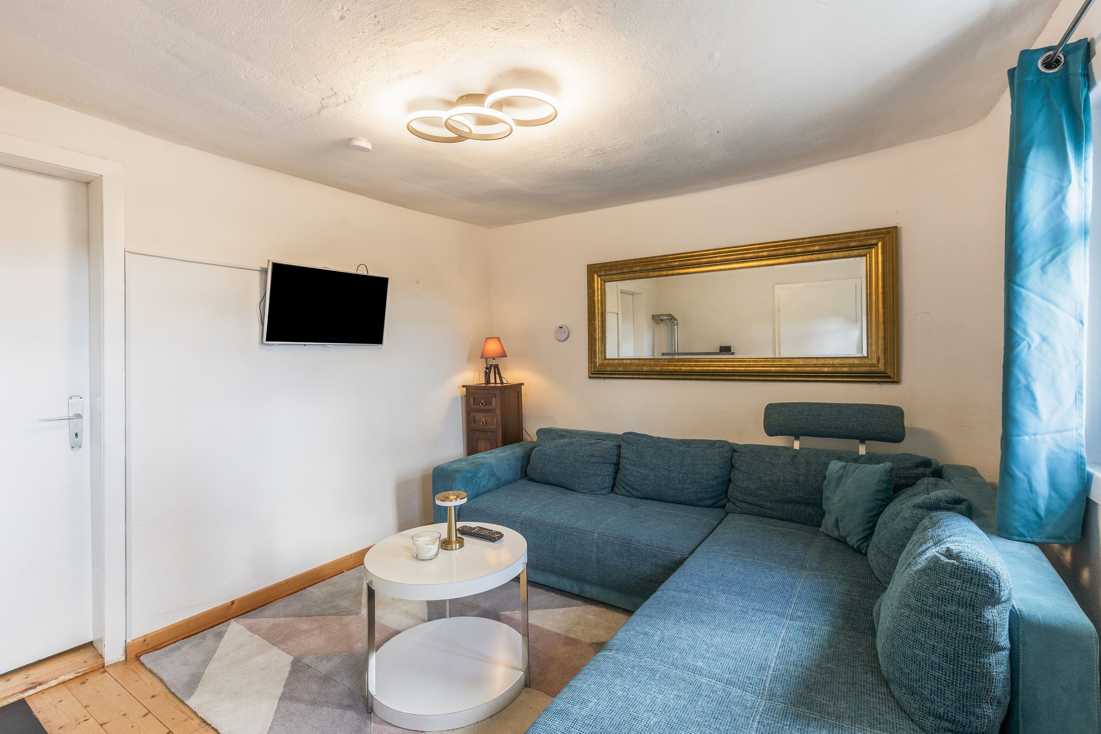 A living room with a sofa, a television mounted on the wall, a small round table with a lamp, and a mirror.