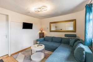 A living room with a sofa, a television mounted on the wall, a small round table with a lamp, and a mirror.