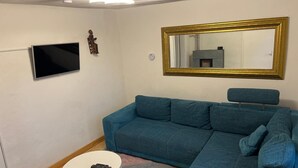 A living room with a teal sofa, a small round table with a remote control, a flat-screen TV, a wall-mounted mirror, and a patterned rug. - Holiday Home 'Ferienhaus Im Grünen Mit Garten' with Private Garden and Wi-Fi (Schotten)