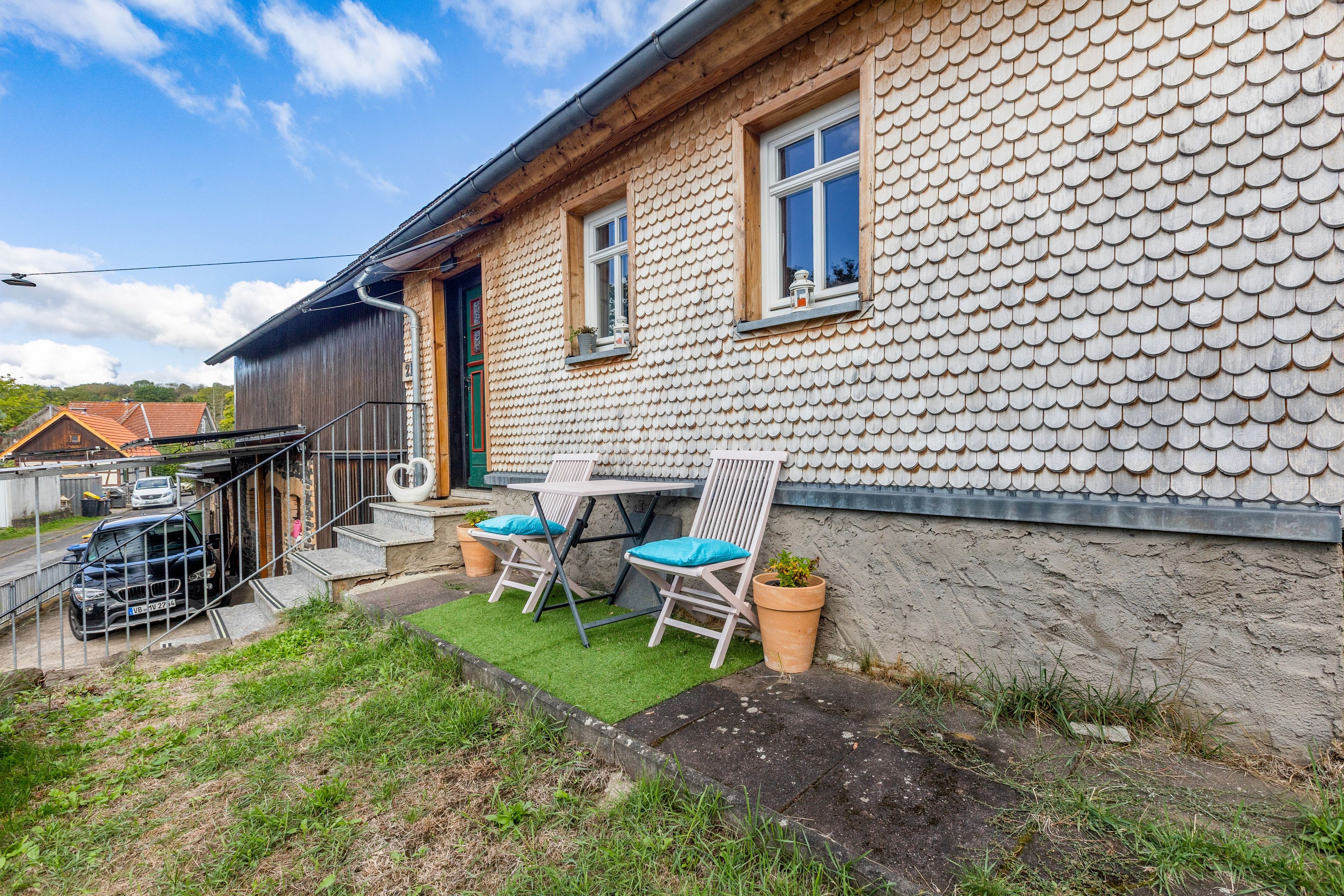 A house with a unique tiled exterior, a small patio with two chairs and a table, and a car parked outside.