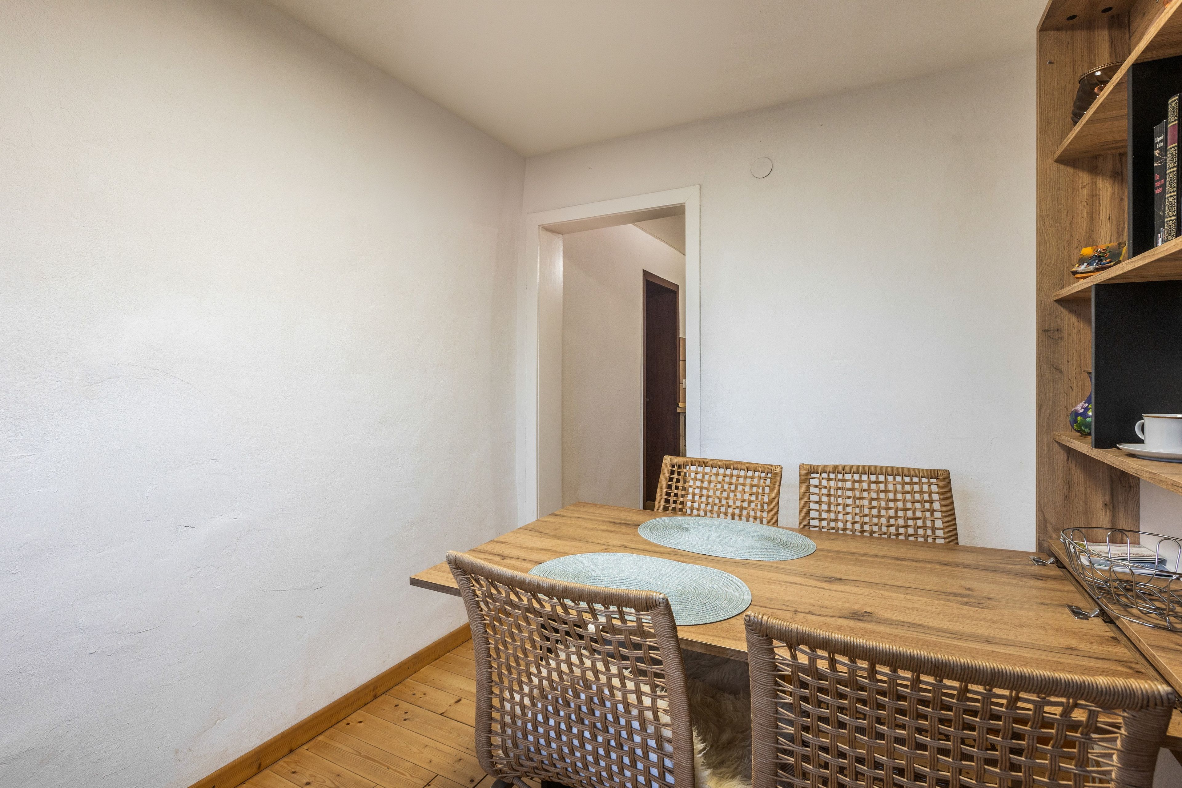 A dining area with a wooden table and wicker chairs, a bookshelf with books and decorative items, and a white door leading to another room.