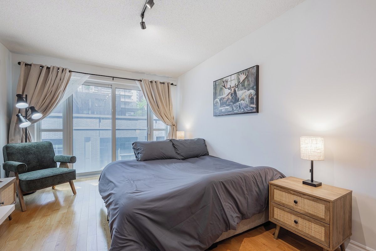 A bedroom with a bed, a chair, a bedside table with a lamp, a window with curtains, and a painting on the wall.