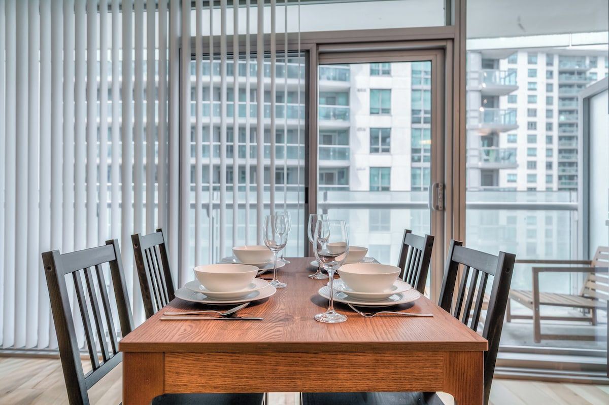 A dining table set with white bowls and wine glasses, surrounded by wooden chairs, in a room with a view of high-rise buildings.