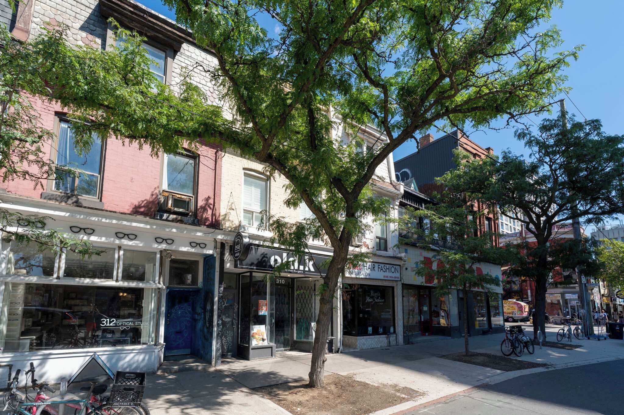 A street lined with multi-story buildings, storefronts, and a tree providing shade.