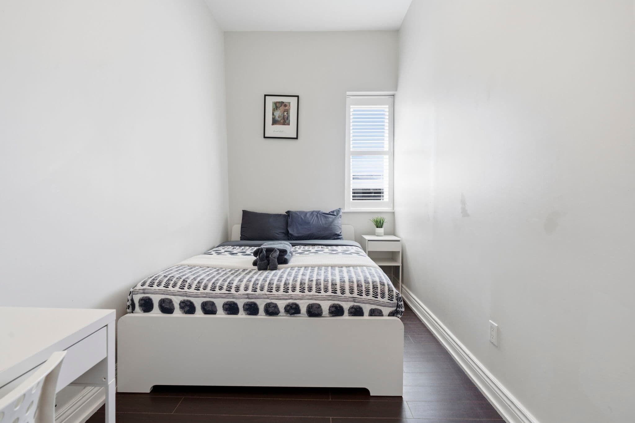 A narrow room with a bed, a small bedside table, a window with blinds, and a wall-mounted picture.