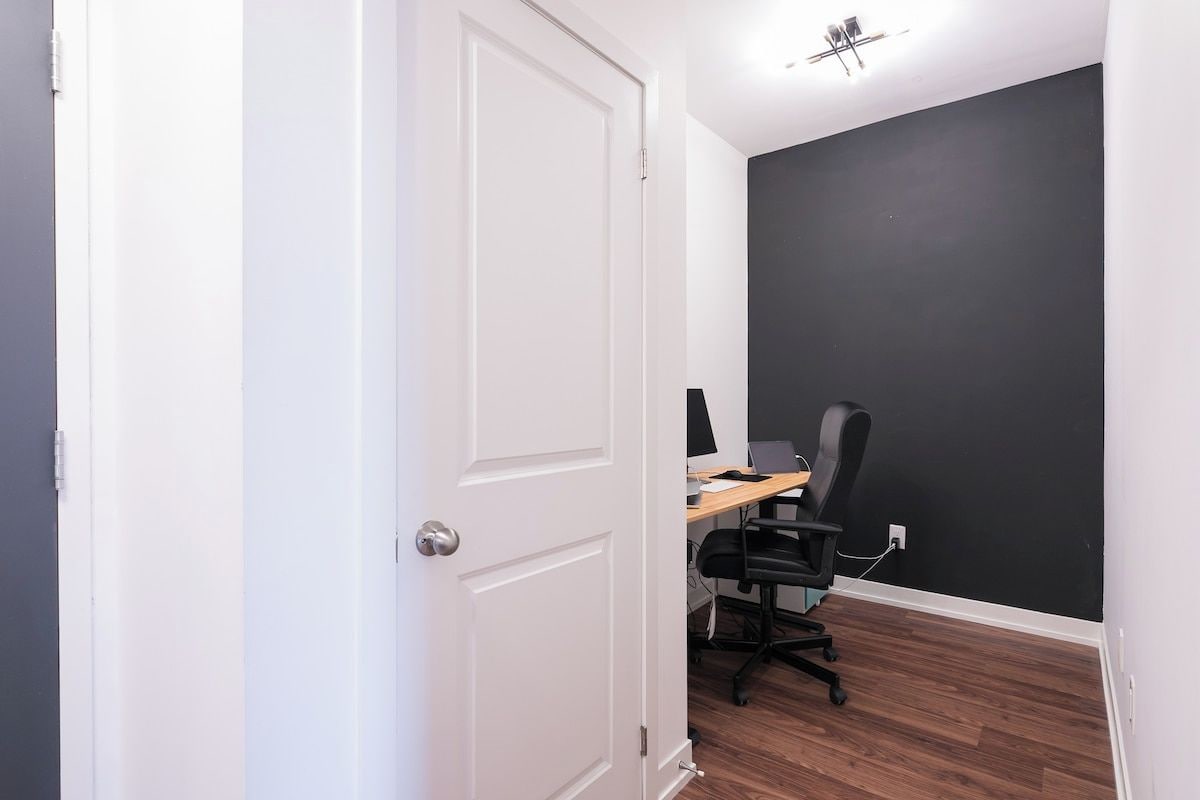 Una habitación con una puerta blanca, un escritorio con una computadora y una silla, y una pared oscura.