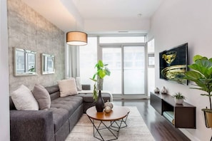 A modern living room with a grey sectional sofa, a round coffee table, and a large window with a view. - Designer Condo 2BR + 1BA Sleeps 5, Near CN Tower (Toronto)