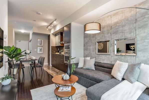 A modern living room with a grey sofa, a dining table with chairs, a coffee table, and a large mirror on the wall. - Designer Condo 2BR + 1BA Sleeps 5, Near CN Tower (Toronto)