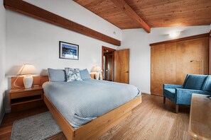A bedroom with a wooden bed, two bedside tables with lamps, a blue chair, a wardrobe, and a framed picture on the wall. - Ocean Song Main | Peak Ocean View, Bluff Trail (Sea Ranch)