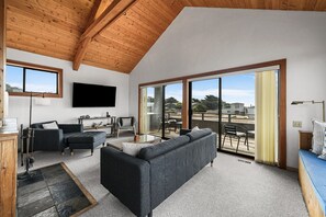 A modern living room with a large TV, comfortable seating, and a balcony with a view. - Ocean Song Main | Peak Ocean View, Bluff Trail (Sea Ranch)