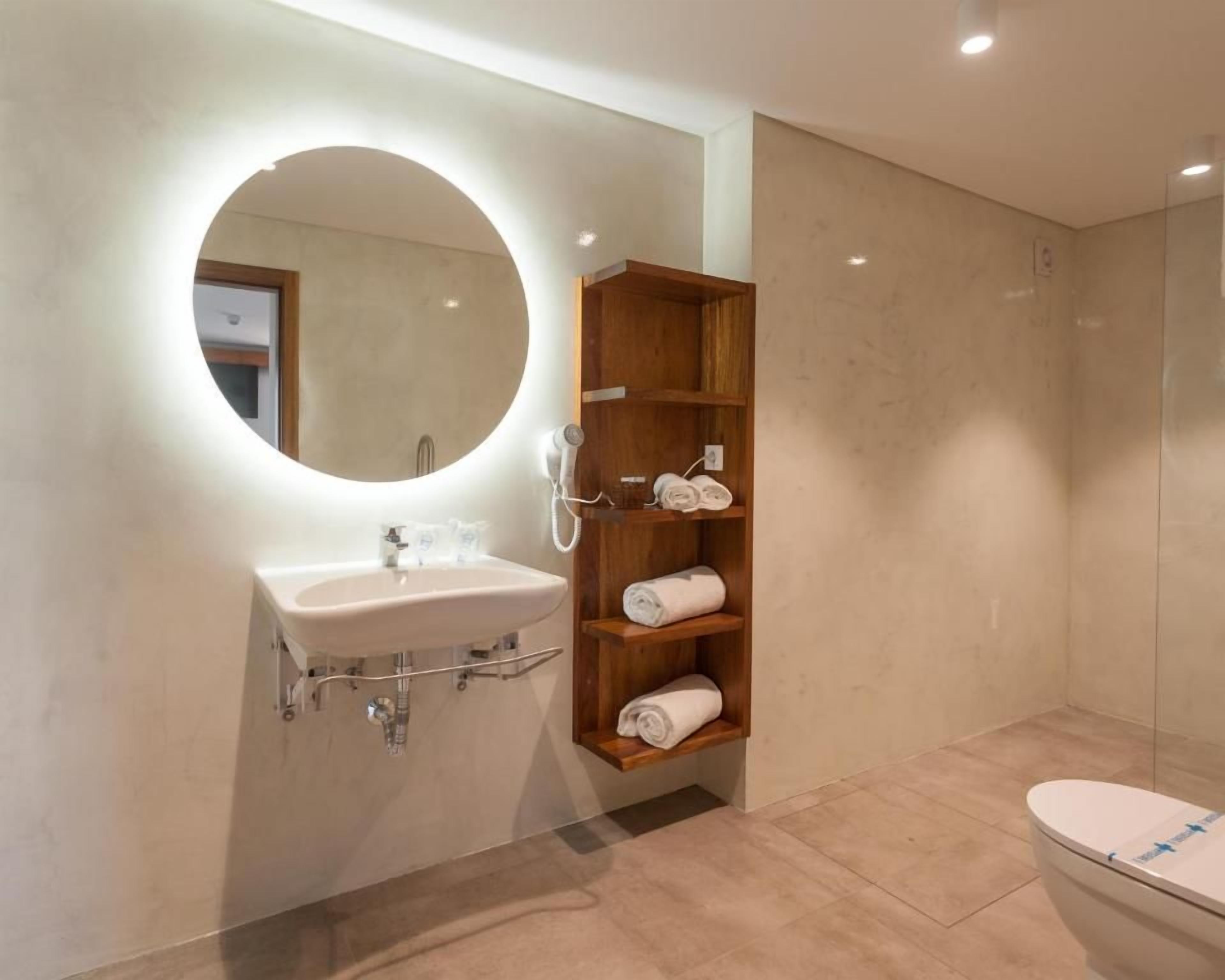 Een moderne badkamer met een ronde spiegel, een wastafel en een toilet. Er is een houten plank met handdoeken.