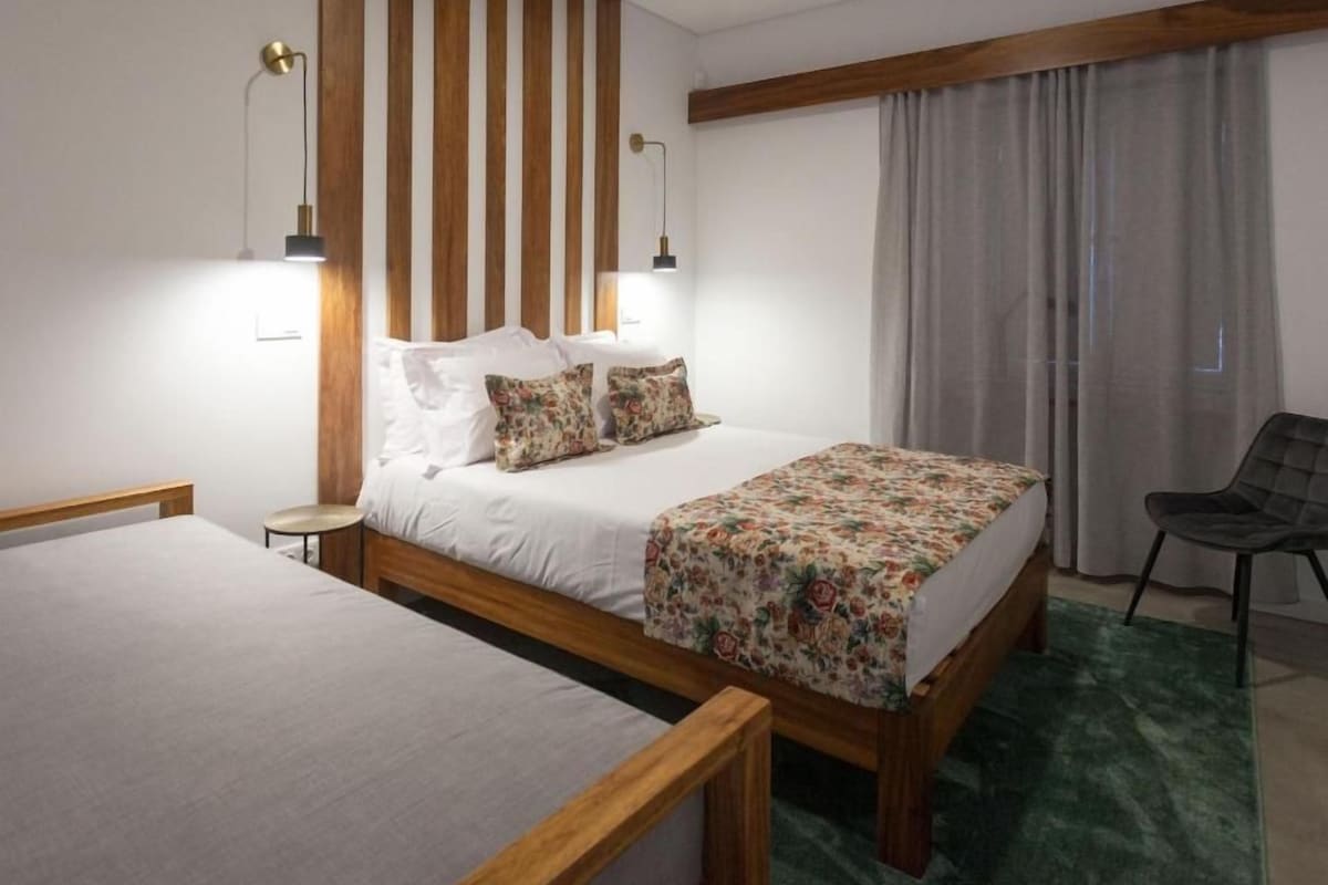 A hotel room with a large bed, a smaller bed, two bedside lamps, a chair, and a window with curtains.