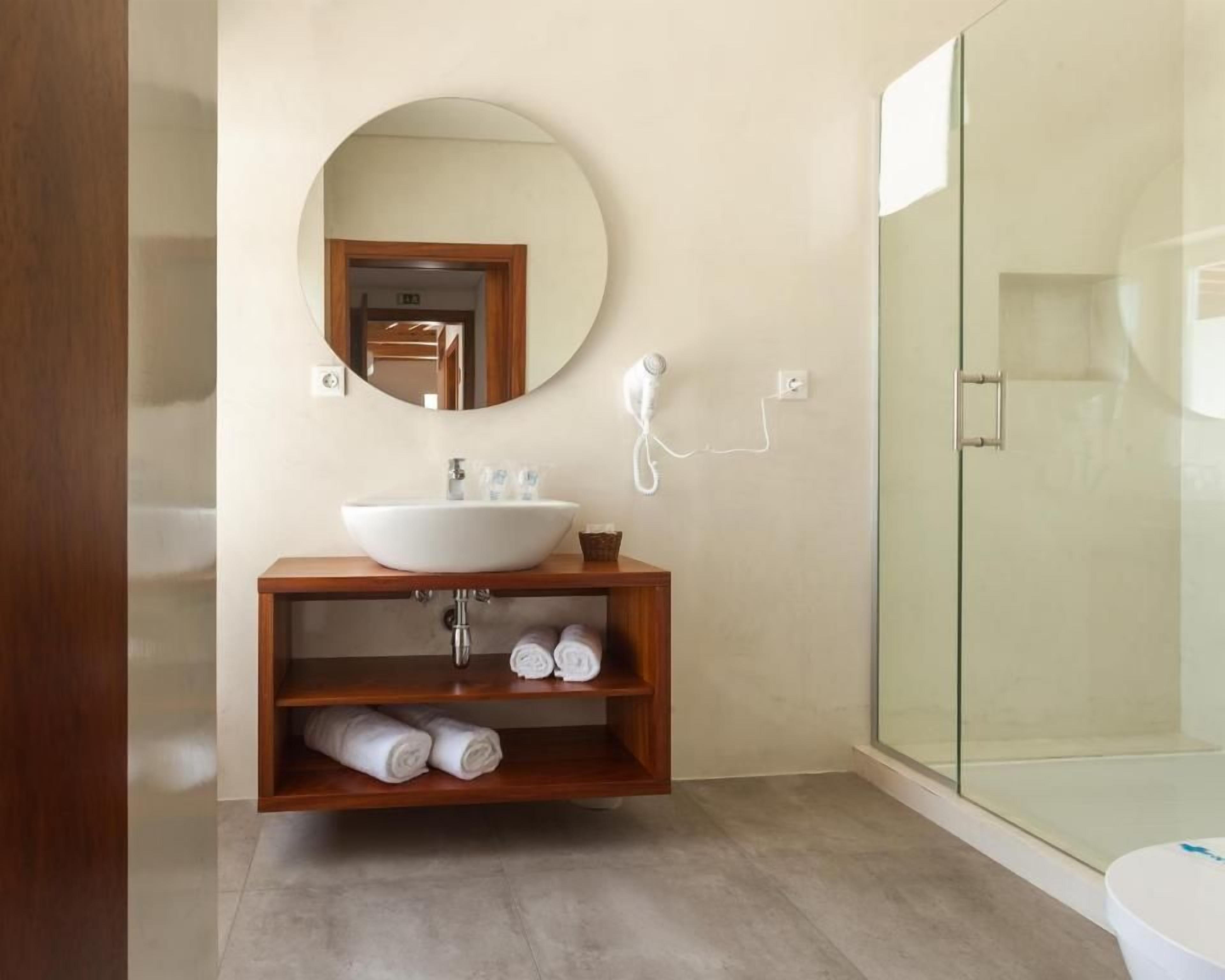 Un baño moderno con un espejo redondo, un tocador de madera, cabina de ducha de vidrio y toallas enrolladas cuidadosamente.