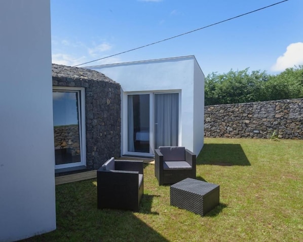 A modern outdoor seating area with a stone wall and a white building with a glass door. - Casa dos Romeiros (SANTA BARBARA)