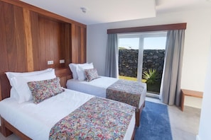 A hotel room with two beds, a large window with a view of a stone wall and plants, and a blue carpet. - Casa dos Romeiros (SANTA BARBARA)
