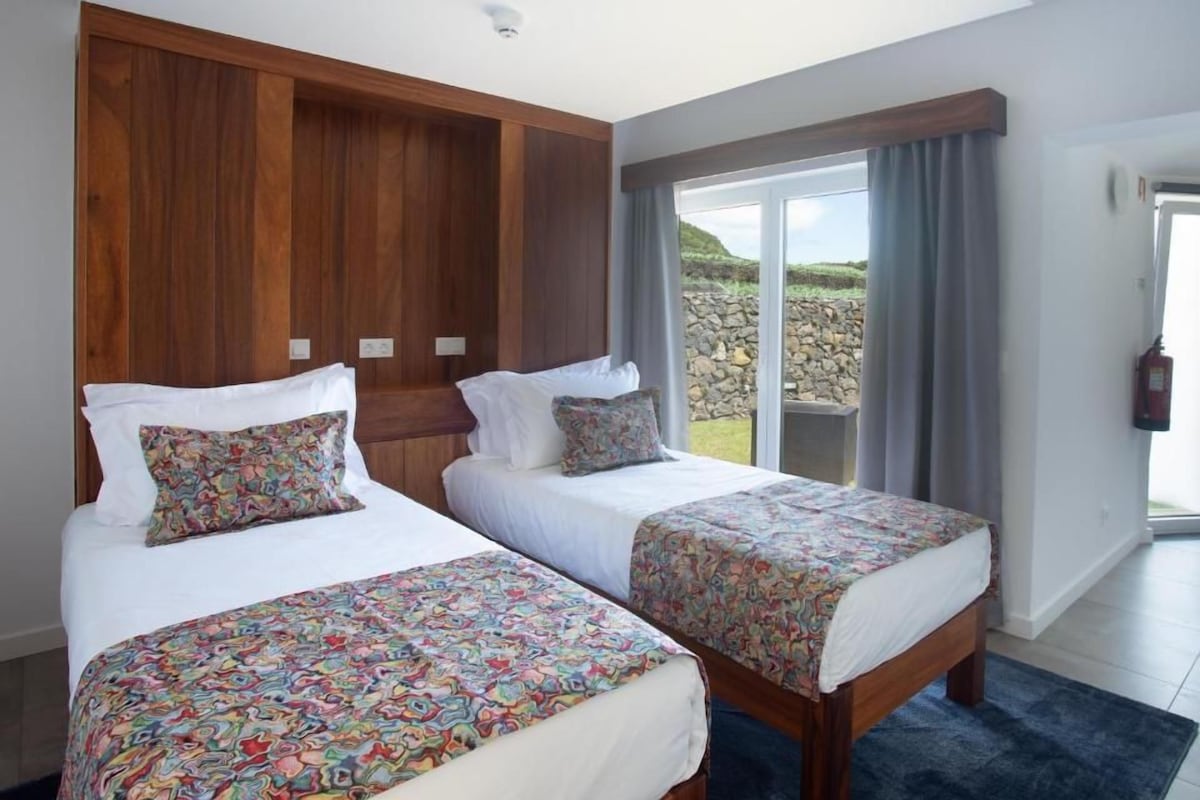A hotel room with two beds, a large window with curtains, a view of the outdoors, and a fire extinguisher on the wall.