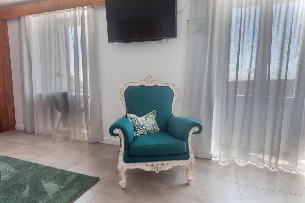 Un sillón verde azulado con un marco decorativo, ubicado en una habitación con alfombra verde, cortinas blancas y un televisor montado en la pared.