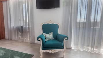 Un sillón verde azulado con un marco decorativo, ubicado en una habitación con alfombra verde, cortinas blancas y un televisor montado en la pared.