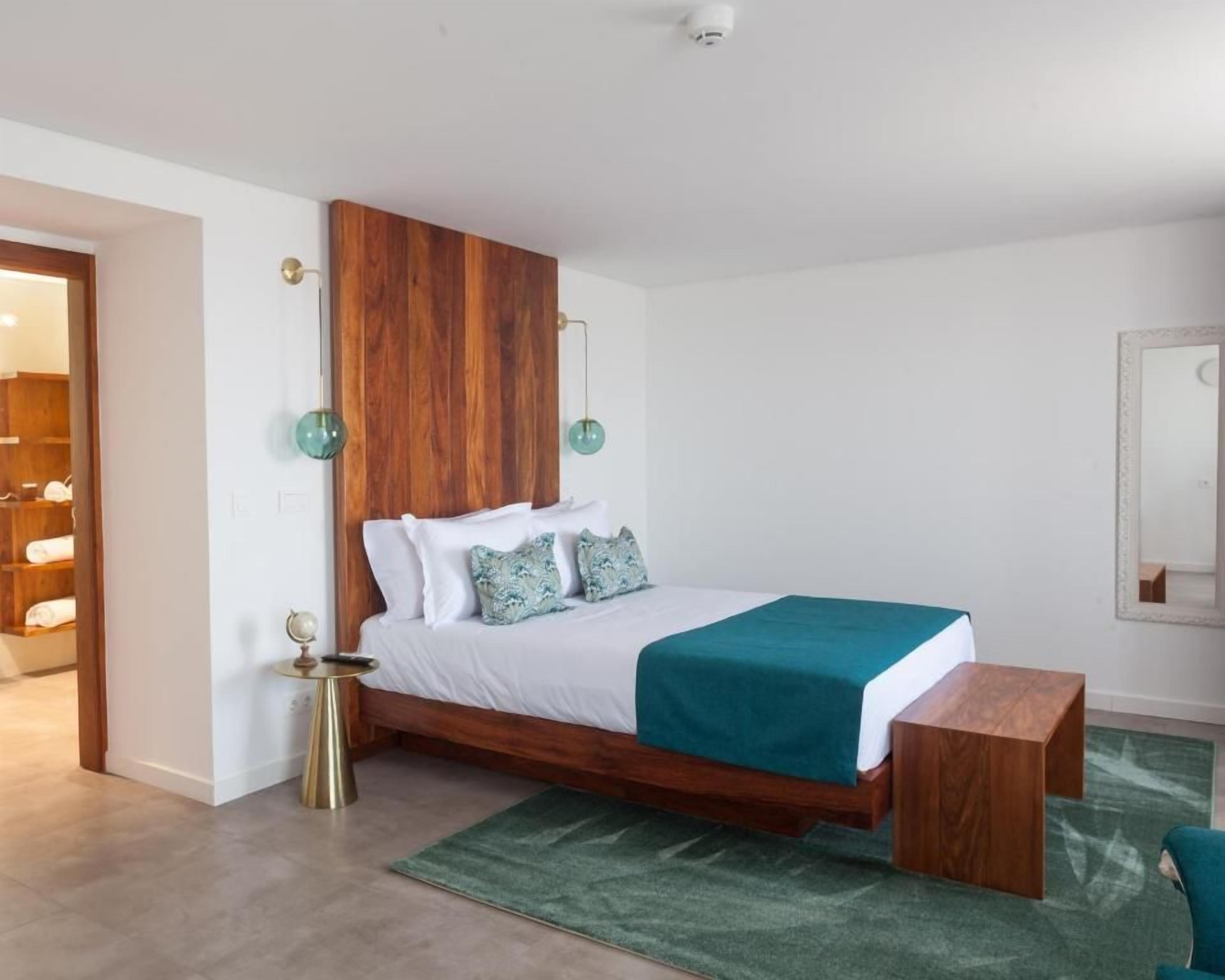 Une chambre avec une tête de lit en bois, un lit avec une literie blanche et un plaid turquoise, une table de chevet sur laquelle se trouve un globe terrestre, et un tapis vert.