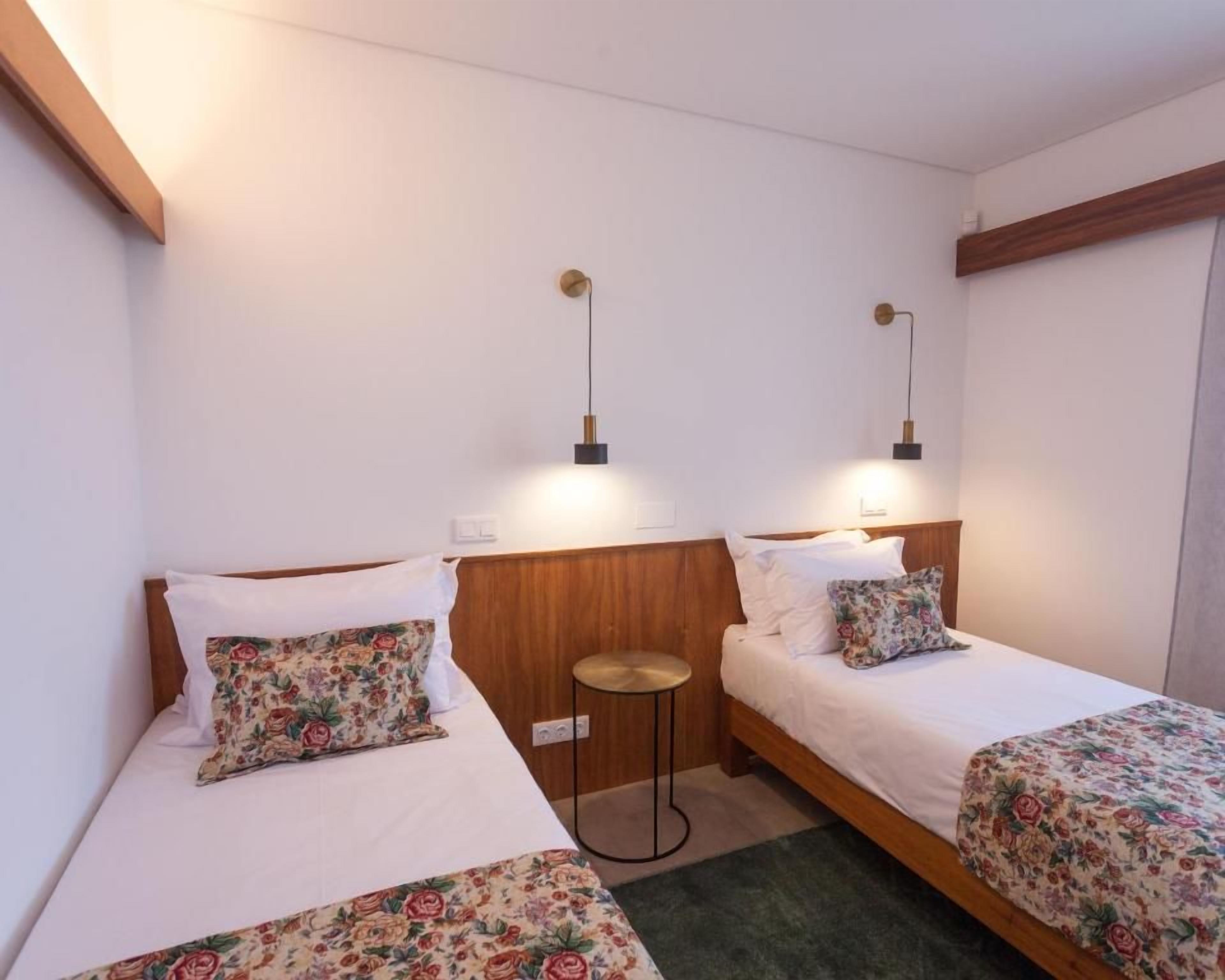 Ein Hotelzimmer mit zwei Betten, jeweils mit floraler Bettwäsche, einem kleinen runden Tisch und Wandlampen.