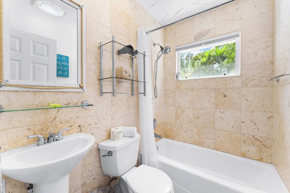 Et badeværelse med badekar, toilet, håndvask, bruser og et vindue med udsigt til grønne omgivelser.