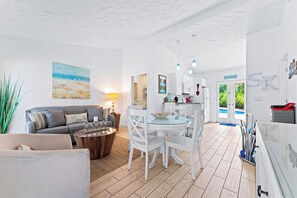 A modern living room with a sofa, coffee table, and dining area with chairs. There is a kitchen visible through an open door. - 2BR villa w/ pool access & beach gear (Siesta Key)