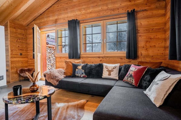 A cozy log cabin interior with a dark gray sofa, wooden walls, and a large window with curtains.