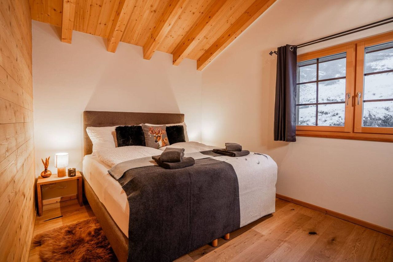 A bedroom with a wooden ceiling, a bed with a grey blanket, a nightstand with a lamp, and a window with curtains.