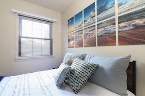 A bedroom with a bed, a window with blinds, and a wall with a series of paintings.