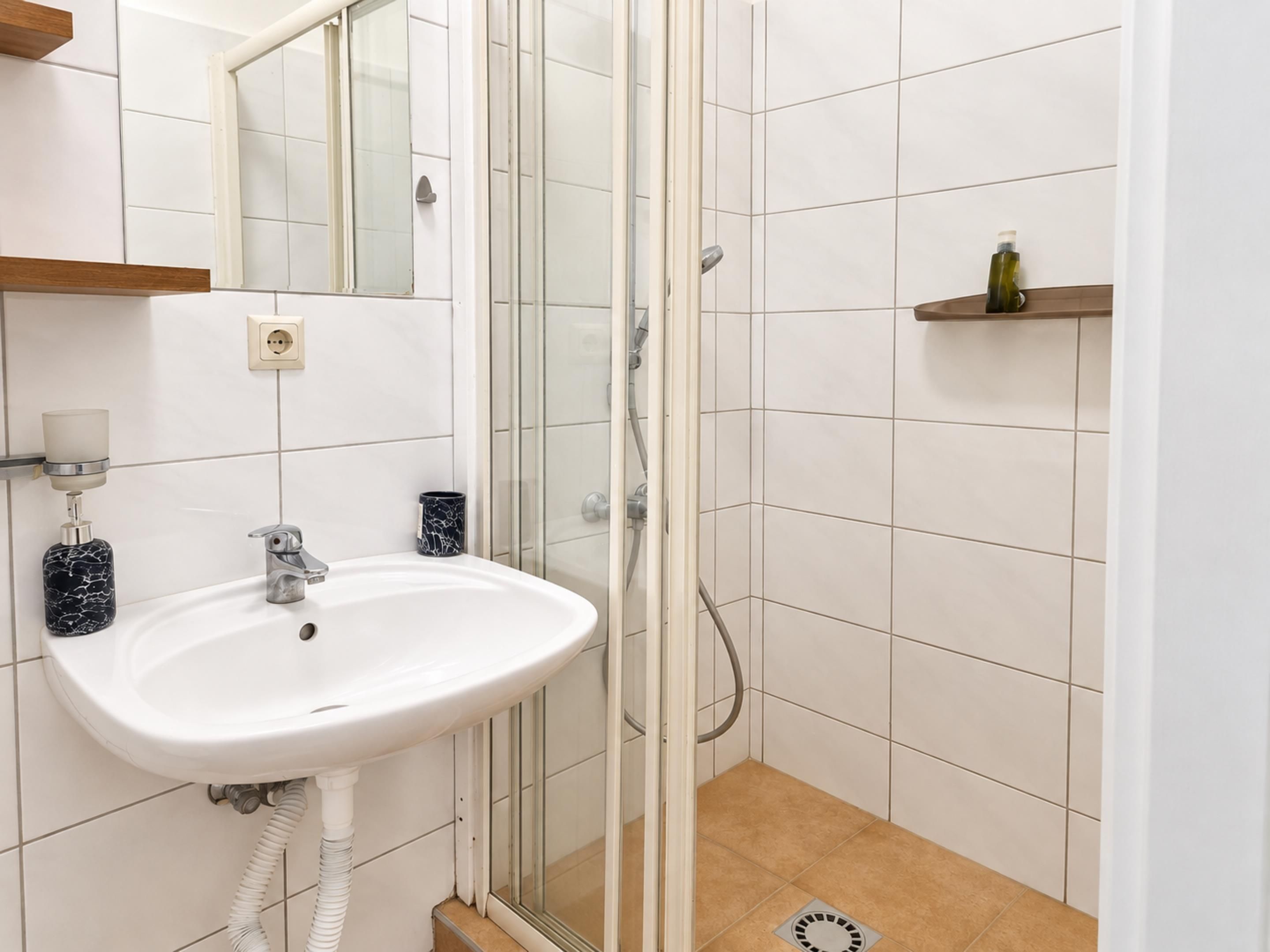 Een badkamer met een wit wastafelmeubel, een douchecabine met glazen wanden en een beigekleurige vloer.