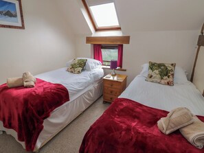 A bedroom with two beds, each with a red blanket and a pillow, a nightstand with a lamp, and a window with curtains. - Chaff House (Worcester)