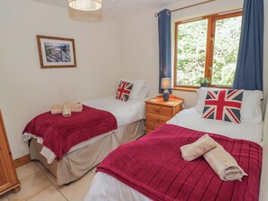 A bedroom with two beds, each with a red blanket and pillows featuring the Union Jack flag. There is a window with blue curtains, a bedside table with a lamp, and a framed picture on the wall. - Kingfisher Lodge (Worcester)