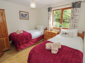 A bedroom with two single beds, a wooden wardrobe, a bedside table with a lamp, and a window with curtains. - Heron Lodge (Worcester)