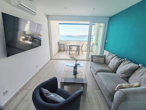 Ein modernes Wohnzimmer mit Fernseher, einer Couch, einem Couchtisch und Blick ins Freie durch eine Schiebetür.