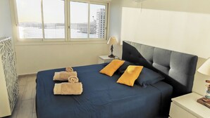 Una habitación de hotel con una cama grande, una ventana con vistas a edificios, una mesita de noche con lámpara y un aire acondicionado de pared.