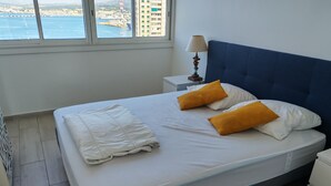 Una cama con ropa de cama blanca y dos almohadas naranjas, una mesita de noche con lámpara y vista al puerto a través de una ventana.