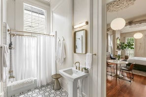 Ein Badezimmer mit Dusche, Waschbecken und Spiegel. Durch einen offenen Durchgang ist ein Essbereich mit Tisch und Stühlen zu sehen.