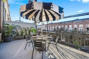 Eine Dachterrasse mit Tisch und Stühlen, einem gestreiften Sonnenschirm und Blumentöpfen.
