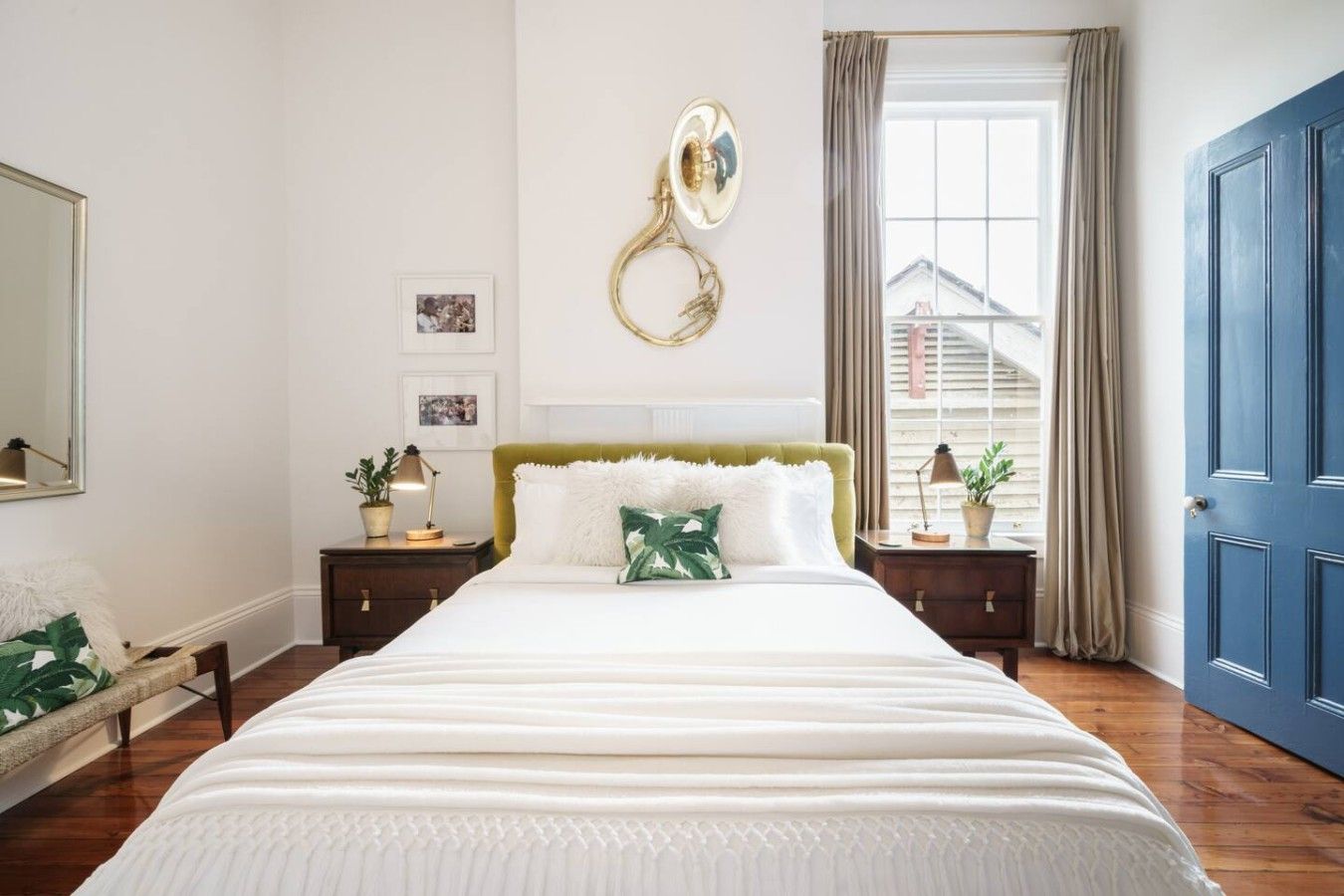 A bedroom with a large bed, a wall-mounted lamp, a window with curtains, and a blue door.