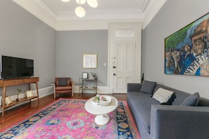 Een woonkamer met een televisie op een houten standaard, een grijze bank, een kamerkleed met dessin en een schilderij aan de muur.