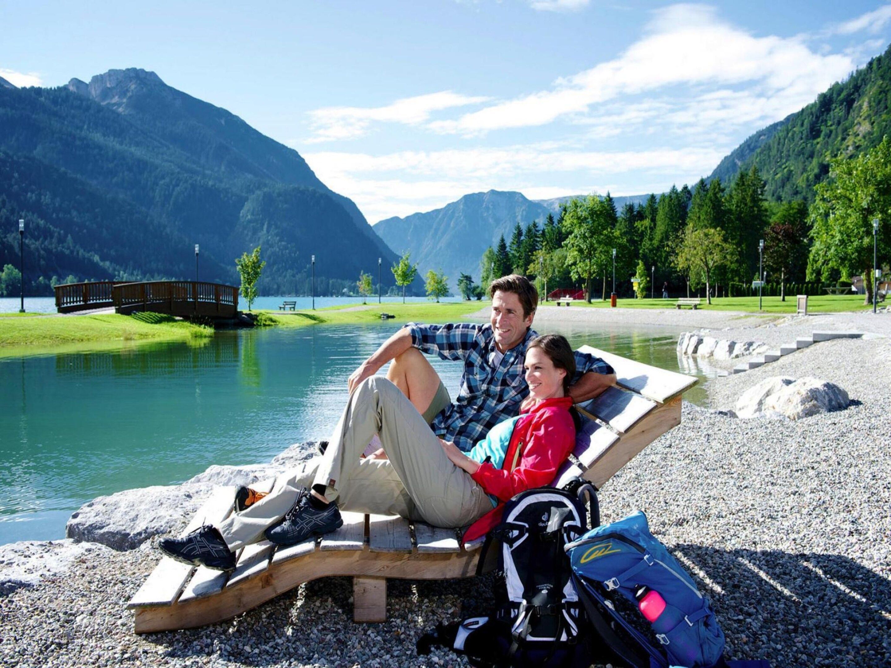 A couple relaxing on a wooden bench by a lake with mountains in the background.