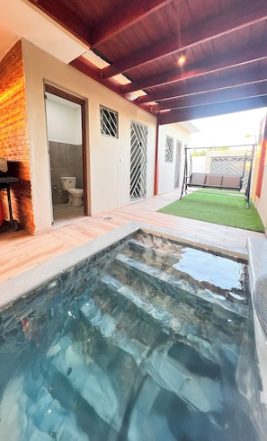 En swimmingpool med fliseterrasse, en murstensvæg, en dør der fører til et badeværelse og et lille græsstykke med en gyngestol.