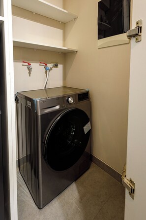 A compact, modern laundry room with a front-loading washer and dryer, a small shelf above the dryer, and a door with a window. - Spacious Suite with a Balcony in Miraflores (Miraflores)